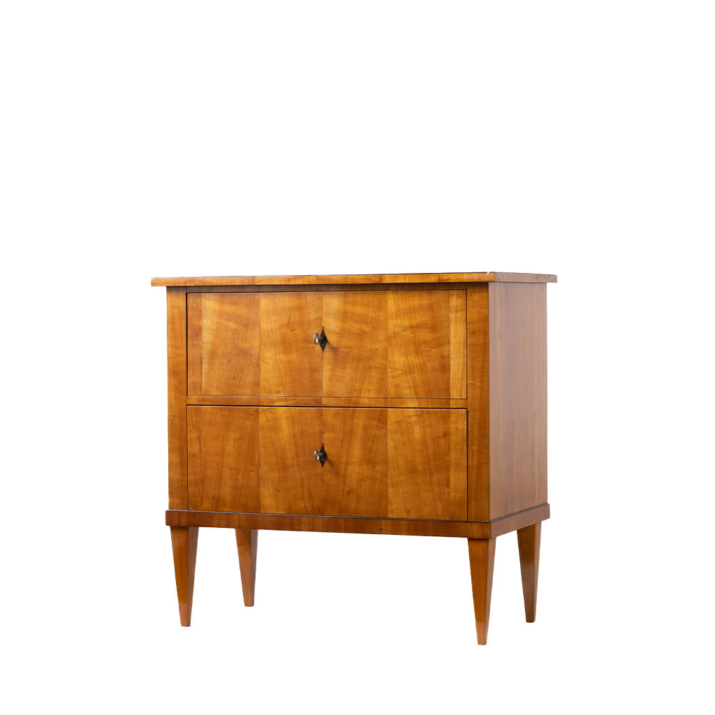 A three-quarter view of an antique Biedermeier two-drawer chest, revealing dovetail joinery and a solid wood interior. The chest features a warm honey-colored wood veneer, simple round pulls, tapered legs, and clean lines that highlight its refined craftsmanship.