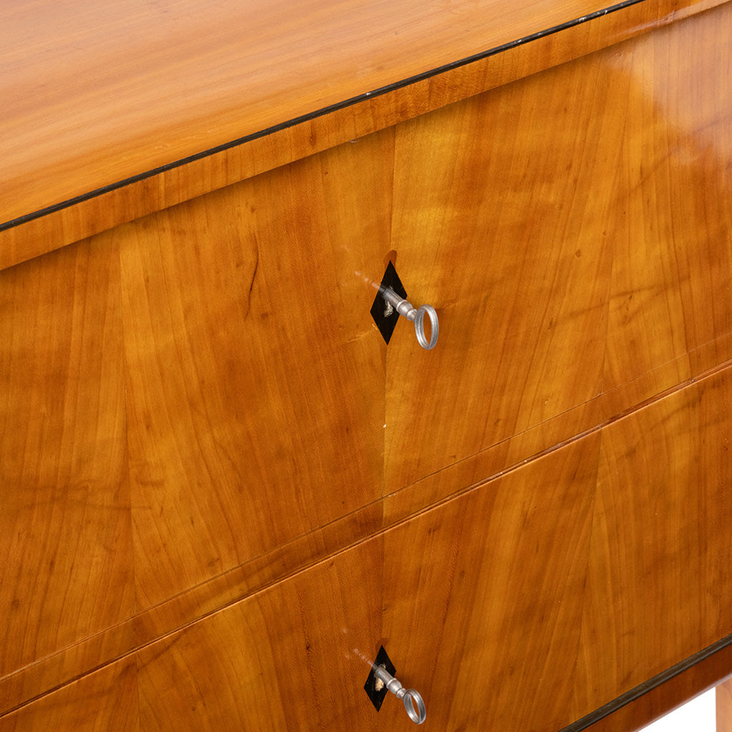 Close-up of a polished wood cabinet or dresser drawer front with warm honey-toned grain, a thin dark edge trim, and silver metal drawer pulls.