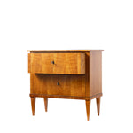 A three-quarter view of an antique Biedermeier two-drawer chest shows the top drawer partially open, revealing dovetail joinery and a solid wood interior. The chest features a warm honey-colored wood veneer, simple round pulls, tapered legs, and clean lines that highlight its refined craftsmanship.
