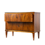 A three-quarter view of an antique Biedermeier two-drawer chest shows the top drawer partially open, revealing dovetail joinery and a solid wood interior. The striking bookmatched walnut veneer, tapered legs, and clean lines highlight the piece's exceptional craftsmanship and elegant design.