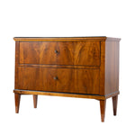 A three-quarter view of an antique Biedermeier two-drawer chest showcases striking bookmatched walnut veneer with dramatic figured wood grain across the drawer fronts. The chest features simple round pulls, tapered legs, and clean, elegant lines characteristic of the Biedermeier style.