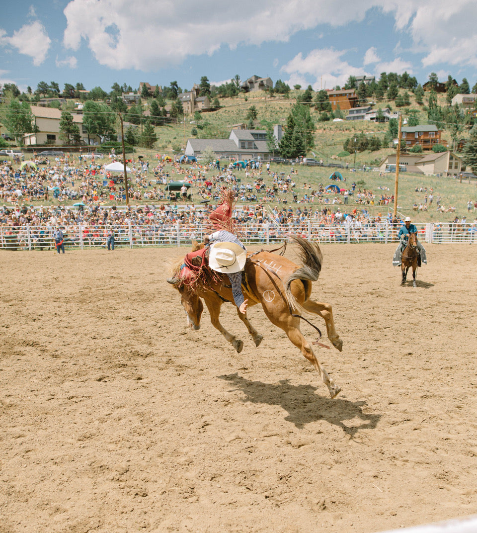 A cowboy rides a bucking horse in a sandy rodeo arena, with a large crowd of spectators and a hillside town in the background under a blue sky with clouds.