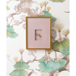 A square framed art piece with a light pink mat and a dragonfly in the center, mounted on a white wallpaper with a botanical pattern.