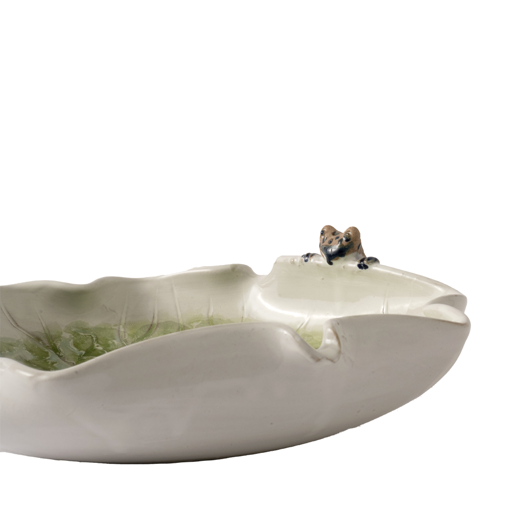 A ceramic leaf-shaped dish with a small brown and black frog figurine perched on the rim, photographed against a plain white background.