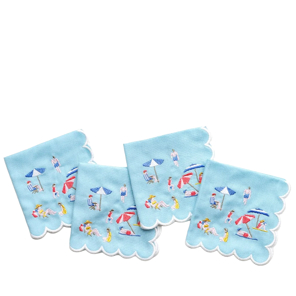Four light blue square napkins with scalloped edges, each printed with a small beach scene featuring people, umbrellas, chairs, and towels, arranged on a white background.