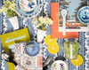 A styled flat-lay collection of home decor and dining accessories, featuring blue and white patterned ceramic plates, a blue teacup and saucer, yellow flowers in white vases, citrus slice coasters, a green Belmond 'Splendido' book, an orange book, and a speckled glass ice bucket.