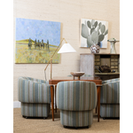 A mid-century modern living room setup featuring four striped armchairs in muted blue, green, and beige tones around a wooden table. The room is styled with a floor lamp, a sideboard cabinet, and two framed wall art prints, including a cactus painting.
