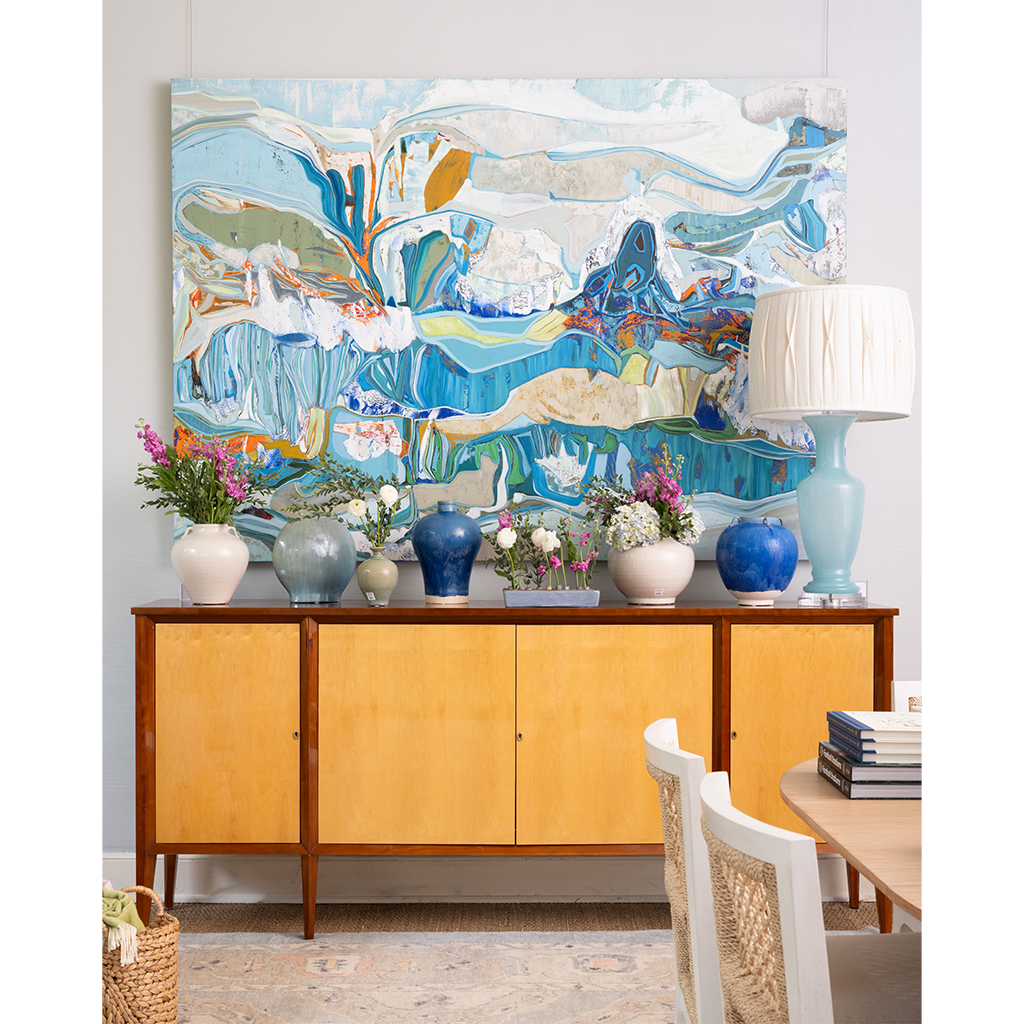 A large abstract canvas painting in shades of blue, white, and beige hangs above a mid-century modern sideboard with yellow wood cabinets. The room is styled with blue and white ceramic vases, potted pink and white flowers, and a light blue table lamp on top.