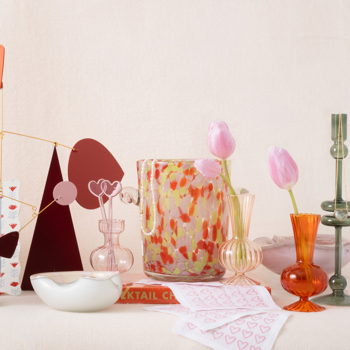 Decorative items including a colorful glass vase, tulips, and a cocktail book on a light surface.