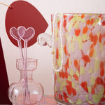 Decorative glass ice bucket with colorful pattern next to a pink hourglass-shaped vase with heart swizzle sticks.