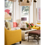 A styled living room interior featuring a mustard yellow armchair, a cream sofa with patterned and coral pillows, a curved cream coffee table, a brown upholstered stool, and floral arrangements by tall windows with beige blinds.