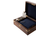 An open dark wood presentation box with brass-tone hardware and a dark blue velvet interior, containing a small clear cut-glass bottle, a silver-toned metal container, and a small key on a white background.