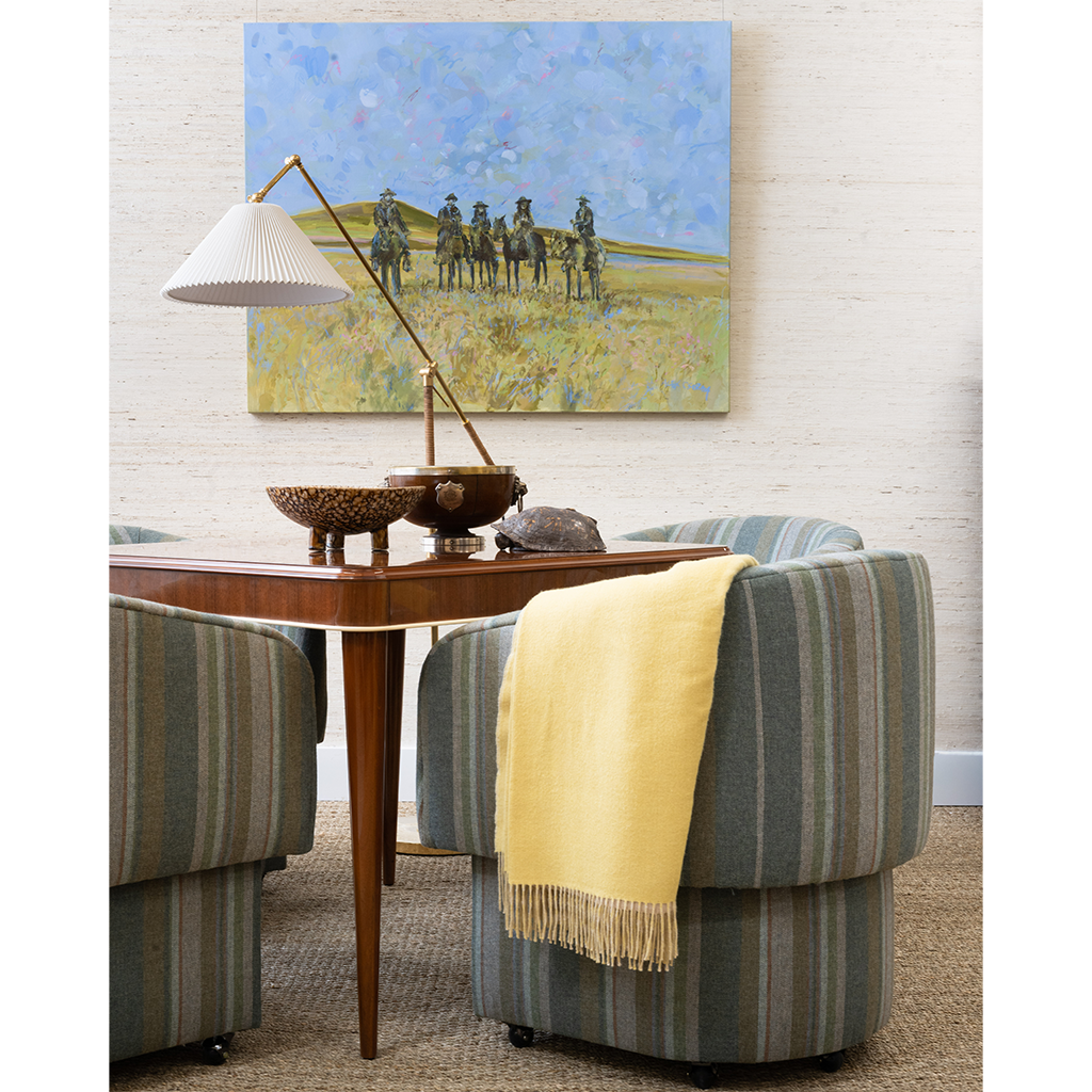 A mid-century modern living room scene featuring a large square canvas painting of soldiers on horseback in a field, hung on a white textured wall above a wooden console table. The room is furnished with striped armchairs in muted green, blue, and beige tones, a yellow throw blanket, and a brass floor lamp.