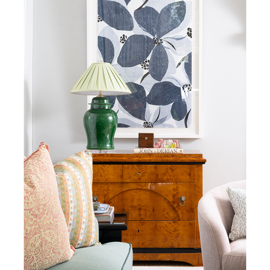A styled interior scene featuring a green ceramic table lamp on a wooden sideboard, with patterned pillows, a light-colored armchair, and a large framed floral artwork on a white wall.