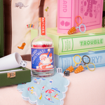 A playful tabletop vignette featuring colorful vintage-style board games, dice, miniature scissors, and a glass bottle filled with matches featuring a festive Santa label. Embroidered textiles, a green game box, and bright pastel accents add a whimsical touch.