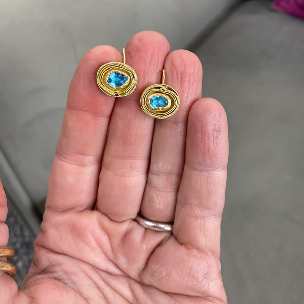 Gold earrings with blue gemstones held in a hand against a neutral background