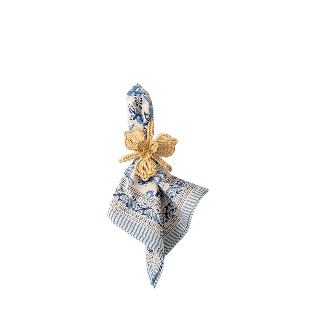 A folded patterned napkin in cream, blue, and white with a gold flower-shaped napkin ring, shown on a plain white background.