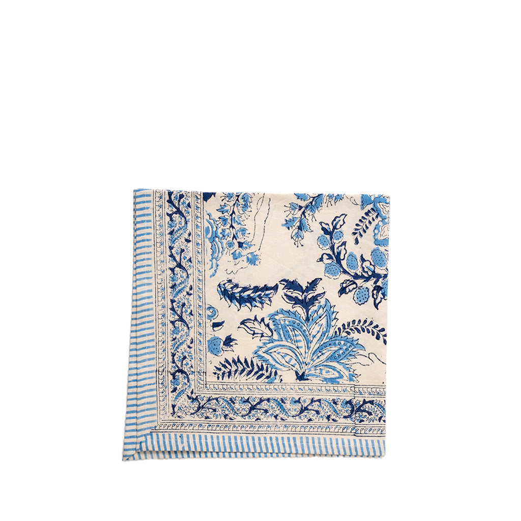 A folded cream-colored cotton napkin with a blue floral and botanical print, featuring a striped border on the left side, set against a plain white background.