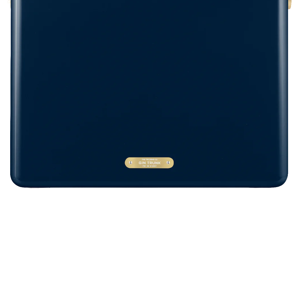 A close-up of the lower front of a navy blue FIELDBAR cooler highlights its smooth, rounded design and a small gold nameplate centered near the base. The gold plaque is engraved with the model name "Gin Trunk" and "Est. MMXXI," adding a refined finishing detail.