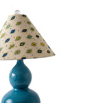 A close-up of a table lamp with a glossy teal blue ceramic base and a conical fabric shade featuring a beige background with blue and yellow geometric patterns, set against a plain white background.