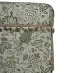 Close-up of upholstered furniture corner showing a botanical floral pattern in muted green and beige, with a beige cord trim and pom-pom detail along the edge.