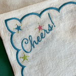 Close-up of a cream-colored linen napkin with a scalloped border, embroidered blue 'Cheers!' script, and three embroidered stars in blue, pink, and green.