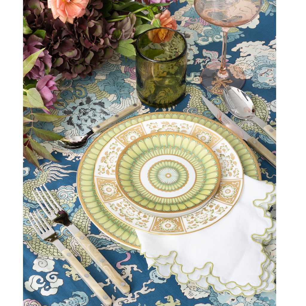 Blue tablecloth with intricate white cloud and mountain patterns under green and gold circular plate settings and floral.