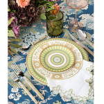 Blue tablecloth with intricate white cloud and mountain patterns under green and gold circular plate settings and floral.