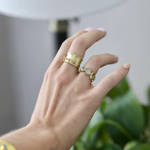 Hand wearing multiple gold rings with gemstones against a blurred indoor background