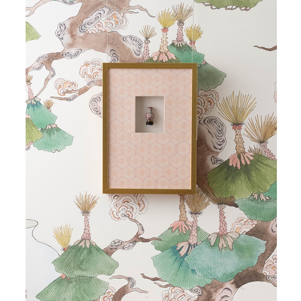A framed wall art piece with a gold frame and a pink patterned backing, mounted on a white wallpapered wall with botanical illustrations.
