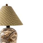A close-up of a table lamp with a beige patterned fabric shade and a ceramic base featuring a black and gold dragon design, set against a plain white background.