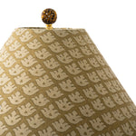 A close-up of a lampshade with a repeating woven floral or leaf-like pattern in beige over a warm olive-brown base, topped with a decorative gold finial.