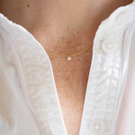 A close-up of a delicate gold chain necklace with a small clear round gemstone pendant, worn on a person's neck over a white button-up shirt.
