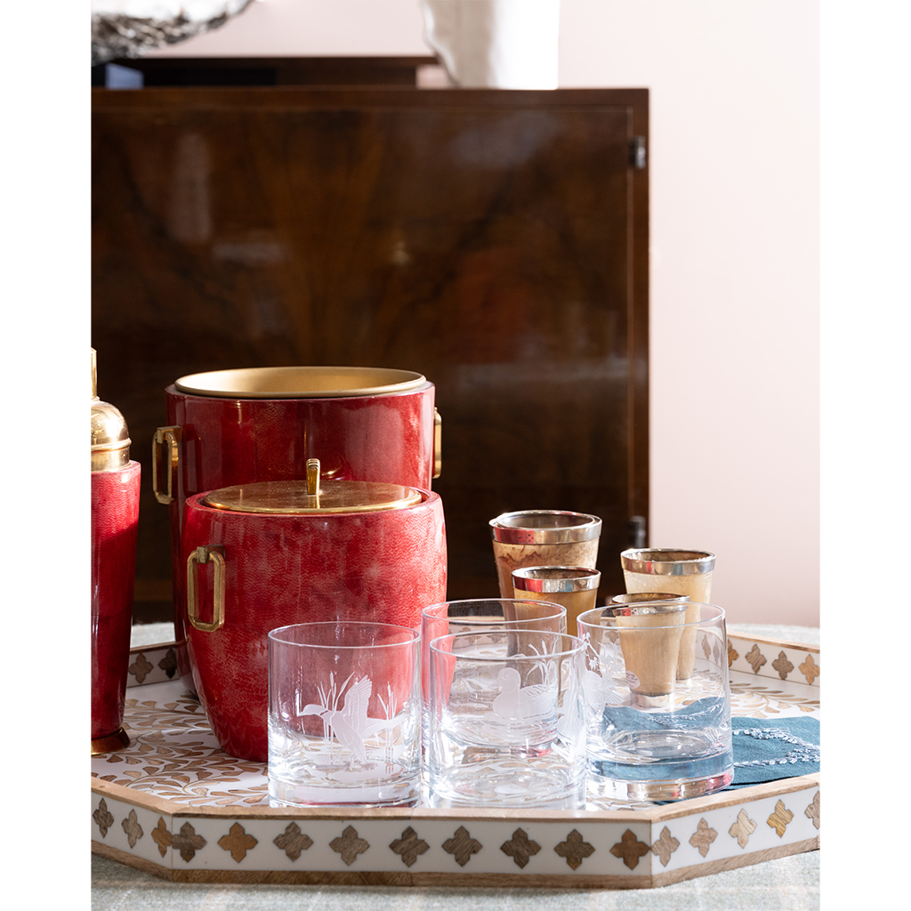 A bar-themed product display featuring a red and gold ice bucket set and matching clear glass tumblers with gold trim, arranged on a decorative tray in an indoor setting.