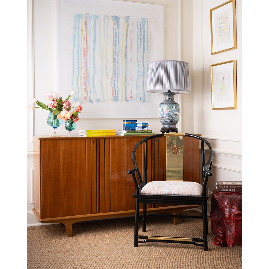 A styled interior scene featuring a mid-century modern wooden credenza with a black wood chair in front, a blue patterned table lamp, and a vase of flowers on top, set against white paneled walls with framed art.