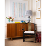 A styled interior scene featuring a mid-century modern wooden credenza with a black wood chair in front, a blue patterned table lamp, and a vase of flowers on top, set against white paneled walls with framed art.