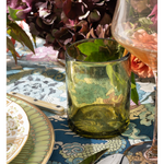 Green glass on a patterned tablecloth with flowers and cutlery in the background