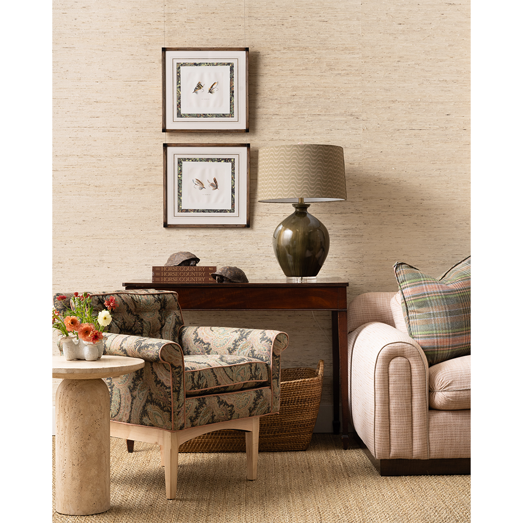 A styled living room corner featuring a beige textured wallpapered wall, a dark wood console table with a green ceramic lamp, two patterned armchairs, and framed bird artwork.