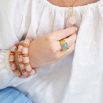 A close-up lifestyle shot of a person wearing a gold ring with a light blue oval stone and a gold necklace with a pink disc charm, styled with peach and white beaded bracelets on a white linen top.