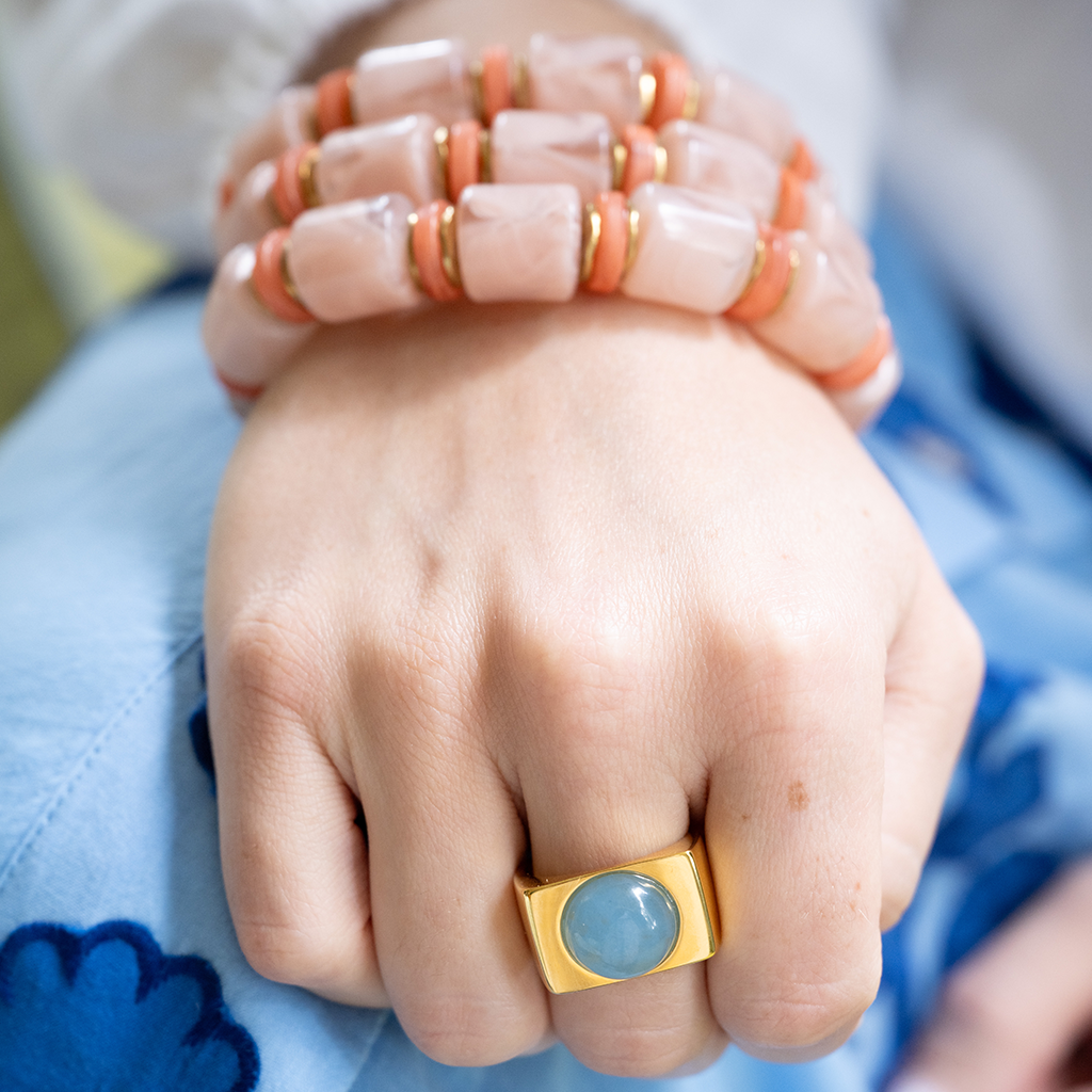 A close-up of a person's hand and wrist wearing a stack of peach and white marbled beaded bracelets with gold-toned spacer rings, along with a gold ring with a blue stone, set against a light blue embroidered garment.