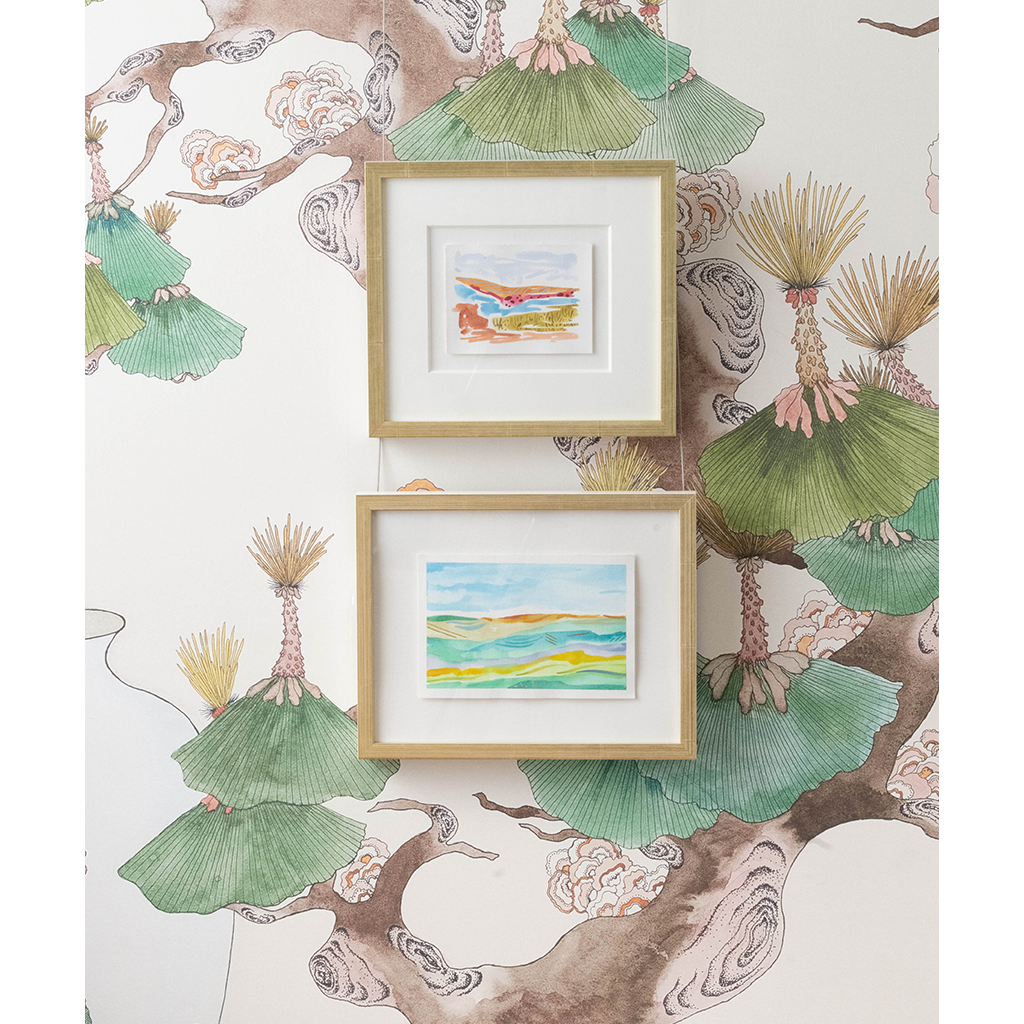 Two small framed landscape paintings hang on a wall with a botanical wallpaper featuring green fan-shaped leaves and brown branches.