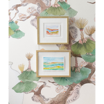 Two small framed landscape paintings hang on a wall with a botanical wallpaper featuring green fan-shaped leaves and brown branches.
