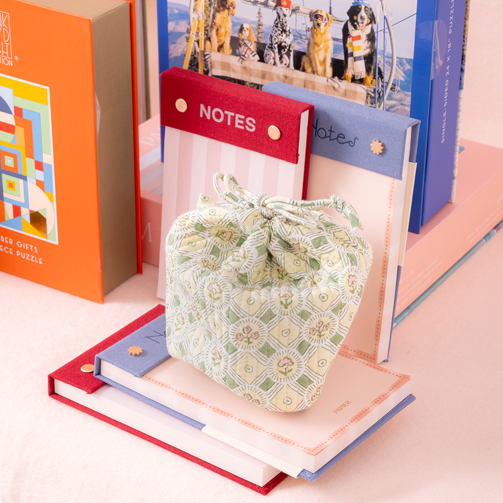 A small drawstring pouch with a sage green and cream geometric pattern sits on top of a stack of pastel-colored books, with stationery items like a red 'NOTES' pad and a blue notebook in the background.