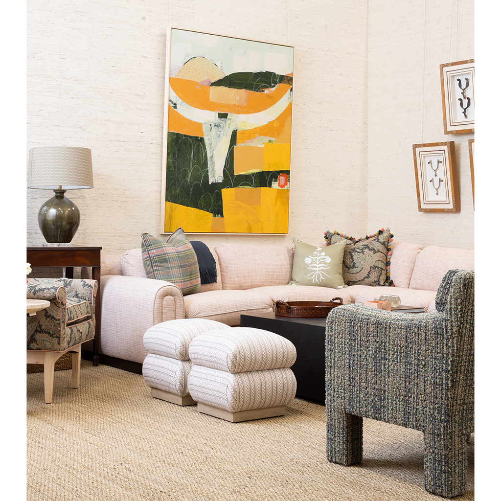 A styled living room corner featuring a light blush pink sectional sofa, striped cream ottomans, a textured blue-gray armchair, a side table with a lamp, and a large abstract art print on the wall.