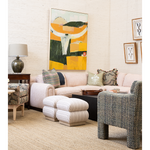 A styled living room corner featuring a light blush pink sectional sofa, striped cream ottomans, a textured blue-gray armchair, a side table with a lamp, and a large abstract art print on the wall.