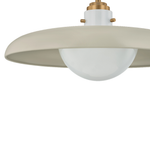 A close-up of a modern pendant light fixture featuring a wide, shallow drum shade in a warm cream-beige finish, a frosted white glass globe bulb, and a brass ceiling mount.