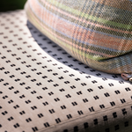 Close-up of a woven outdoor chair cushion in a light beige fabric with a black geometric pattern, with a plaid cushion resting on top.