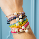 A stack of colorful braided cord bracelets with white faux pearls and gold-toned hardware, worn on a person's wrist against a soft blue paneled background.