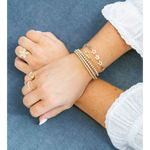 A close-up of crossed hands and forearms wearing delicate gold jewelry, including a flower-shaped ring, a small band ring, and several stacked bracelets, against a textured blue fabric background.