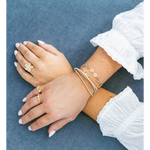 A close-up of crossed hands and forearms wearing delicate gold jewelry, including a flower-shaped ring, a small band ring, and several stacked bracelets, against a textured blue fabric background.